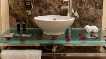 A bathroom with a marble wall, a white vessel sink, and a glass shelf with toiletries and rose petals.