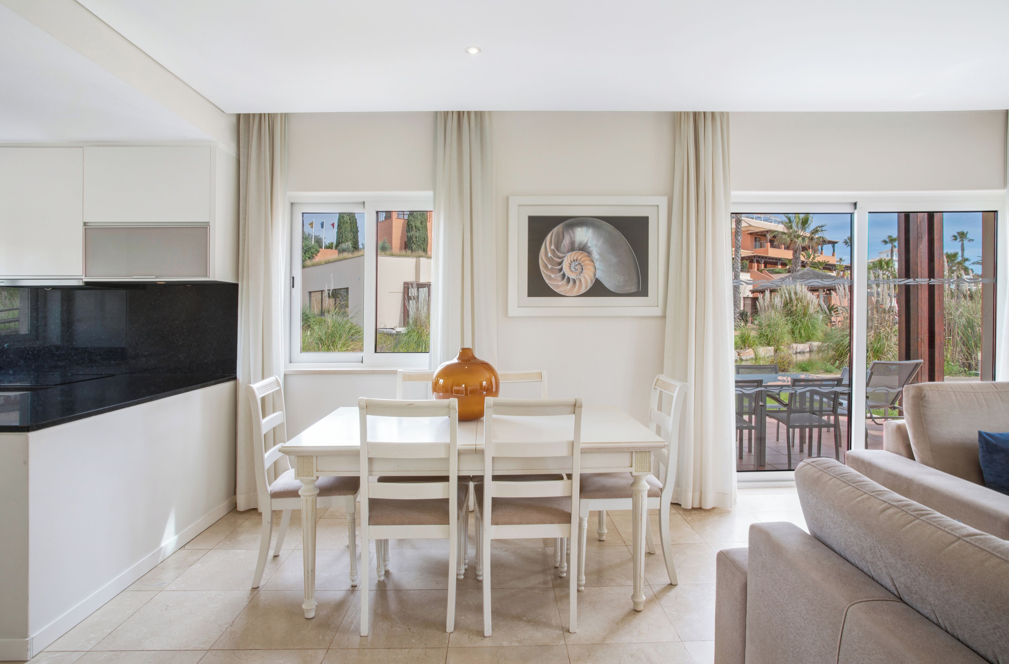 A modern kitchen with white cabinets, a black countertop, and a dining area with a white table and chairs. There is a large window with a view of buildings and greenery.