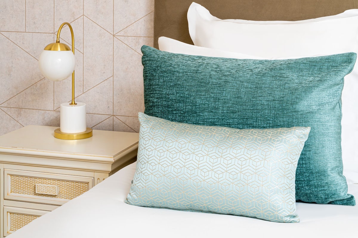 A neatly arranged bedroom with a geometric-patterned wall, a bedside table with a lamp, and a bed with multiple pillows.