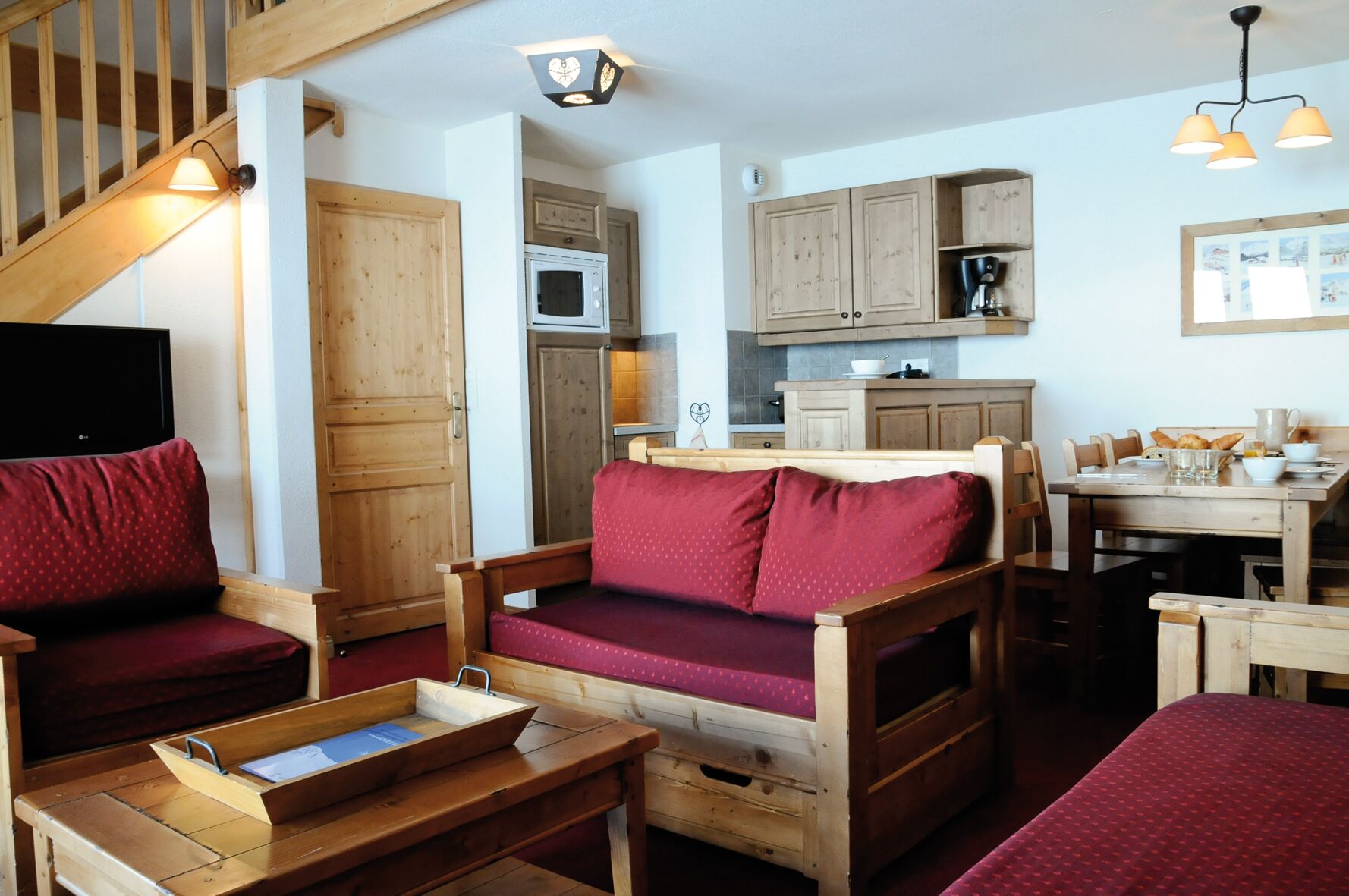 A cozy room with a wooden staircase, a sofa with red cushions, a wooden dining table, and a kitchen area with wooden cabinets and a microwave.