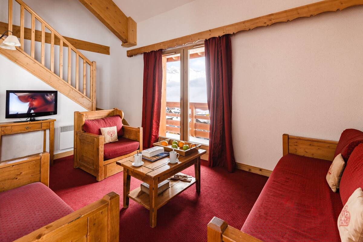 Una habitación acogedora con una escalera de madera, un sofá, una silla, una mesa con libros y fruta, y un televisor.