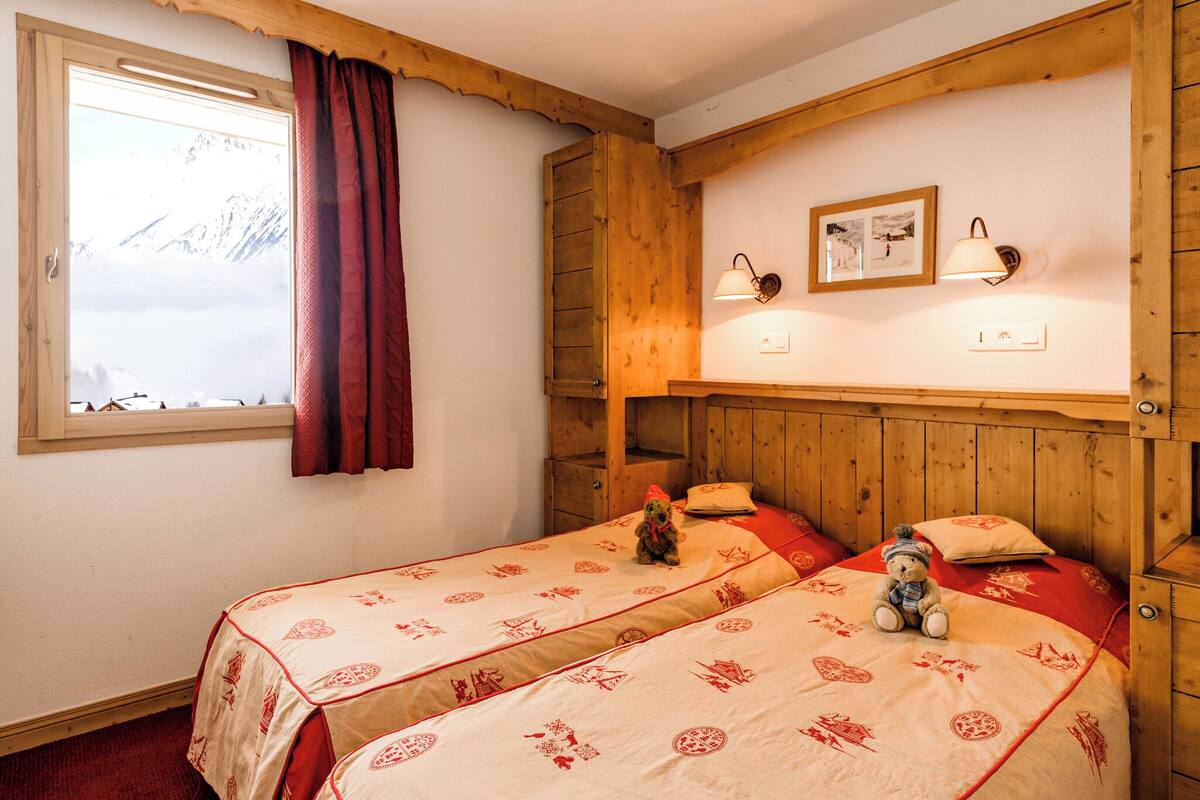 Una habitación con dos camas, cabecero de madera, ventana con vistas a las montañas y un osito de peluche sobre una de las camas.