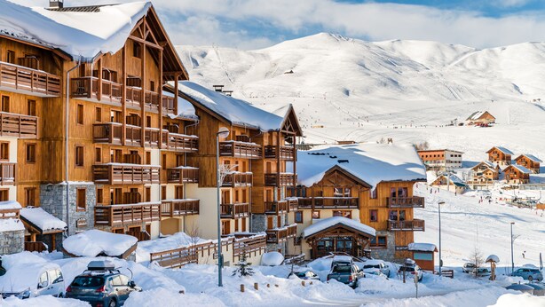 Estación de esquí cubierta de nieve con edificios de madera, incluyendo un albergue de varios pisos con balcones y un telesilla al fondo.
