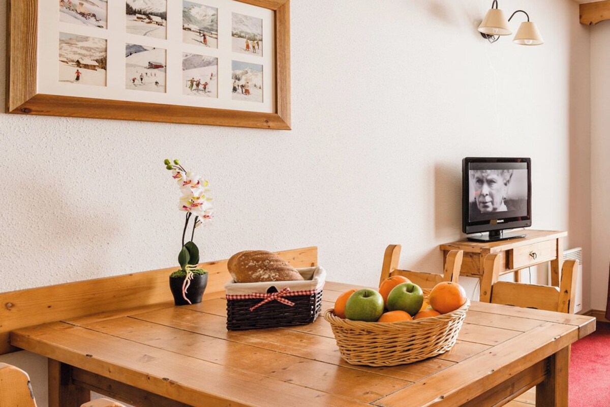 Une table à manger en bois, entourée de chaises, un panier de fruits, un vase rempli de fleurs, un autre panier contenant du pain, et un tableau encadré accroché au mur.