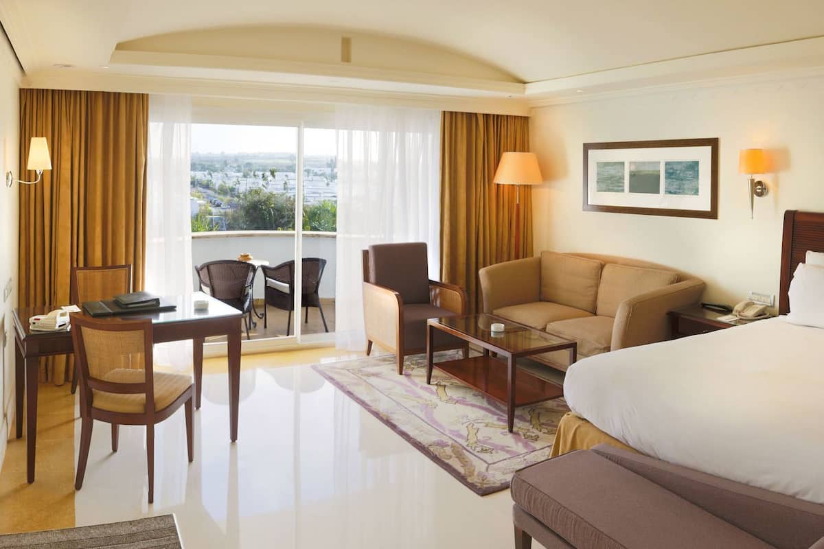 A hotel room with a bed, a desk with a chair, a sofa, a TV, and a balcony with curtains.