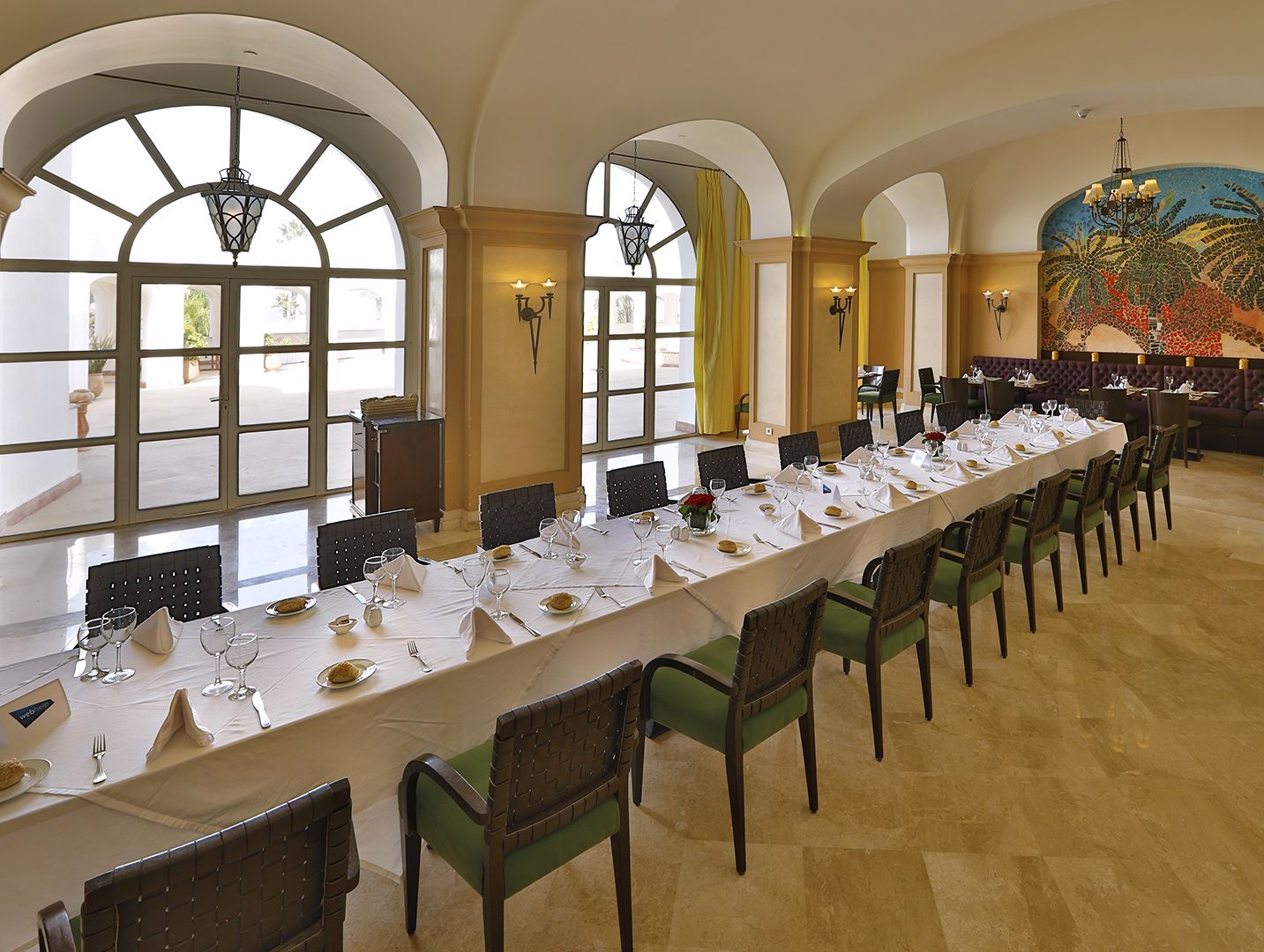 Une longue table à manger, dressée pour un repas, avec des chaises, de grandes fenêtres et une fresque murale.