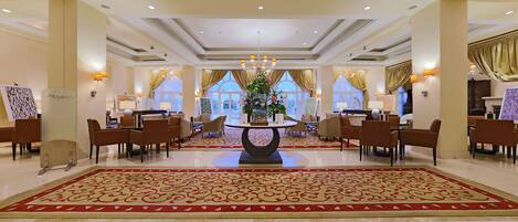 A spacious lobby with a central round table, multiple seating areas, and large windows with curtains.