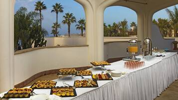 Une table de buffet proposant une variété de pâtisseries et de desserts, un coin café et une vue sur les palmiers à travers de grandes fenêtres en arc de cercle.