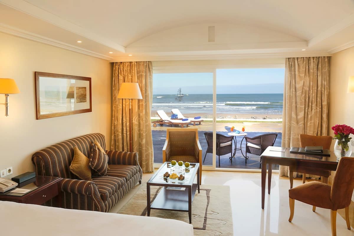 A hotel room with a large bed, a striped sofa, a dining table with chairs, a coffee table, and a view of the beach and ocean.