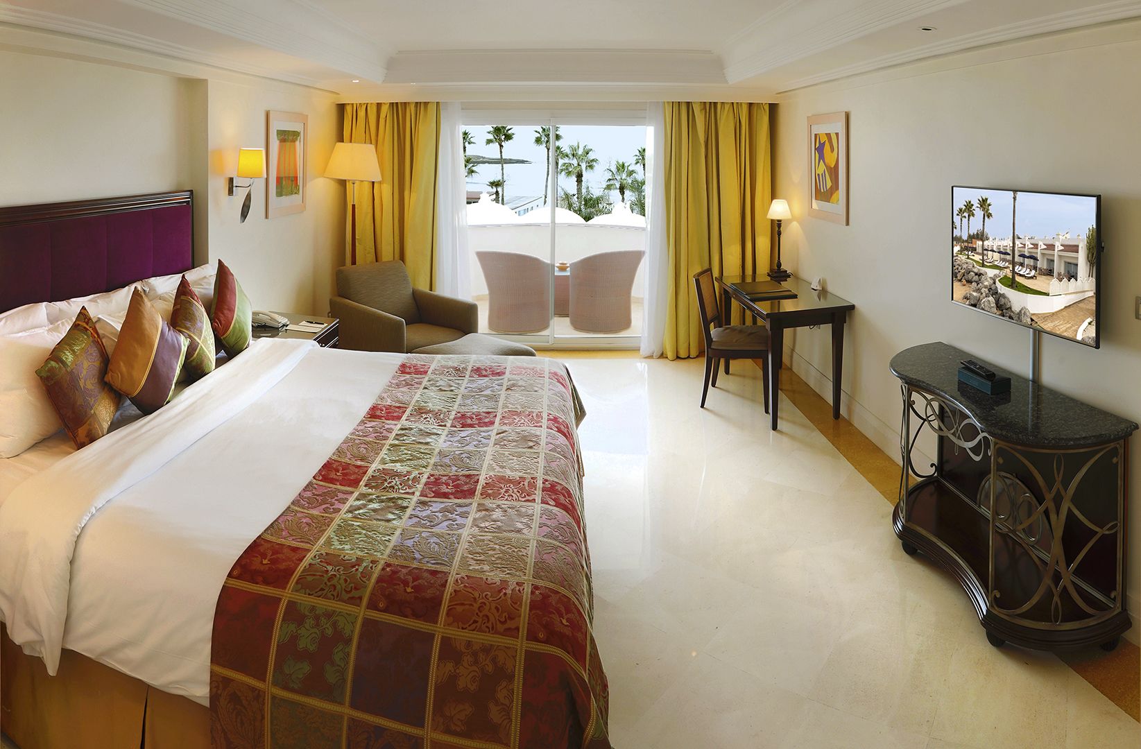 Ein Hotelzimmer mit einem großen Bett, einem Schreibtisch, einem Sessel, einem Fernseher und einem Balkon mit Blick auf Palmen und das Meer.