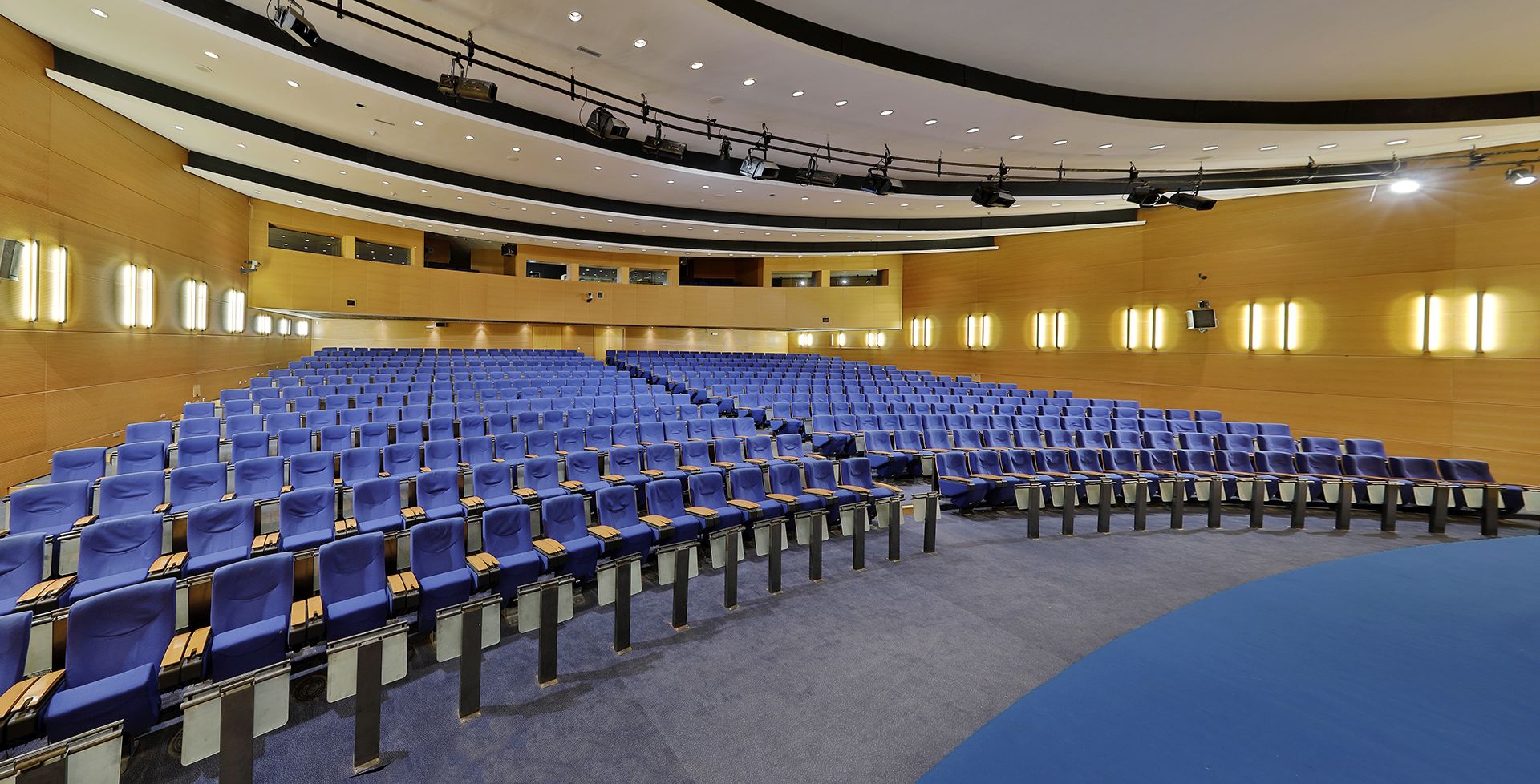 A large auditorium with rows of blue chairs, a stage with lighting equipment, and a curved ceiling.