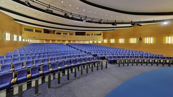 A large auditorium with rows of blue chairs, a stage with lighting equipment, and a curved ceiling.