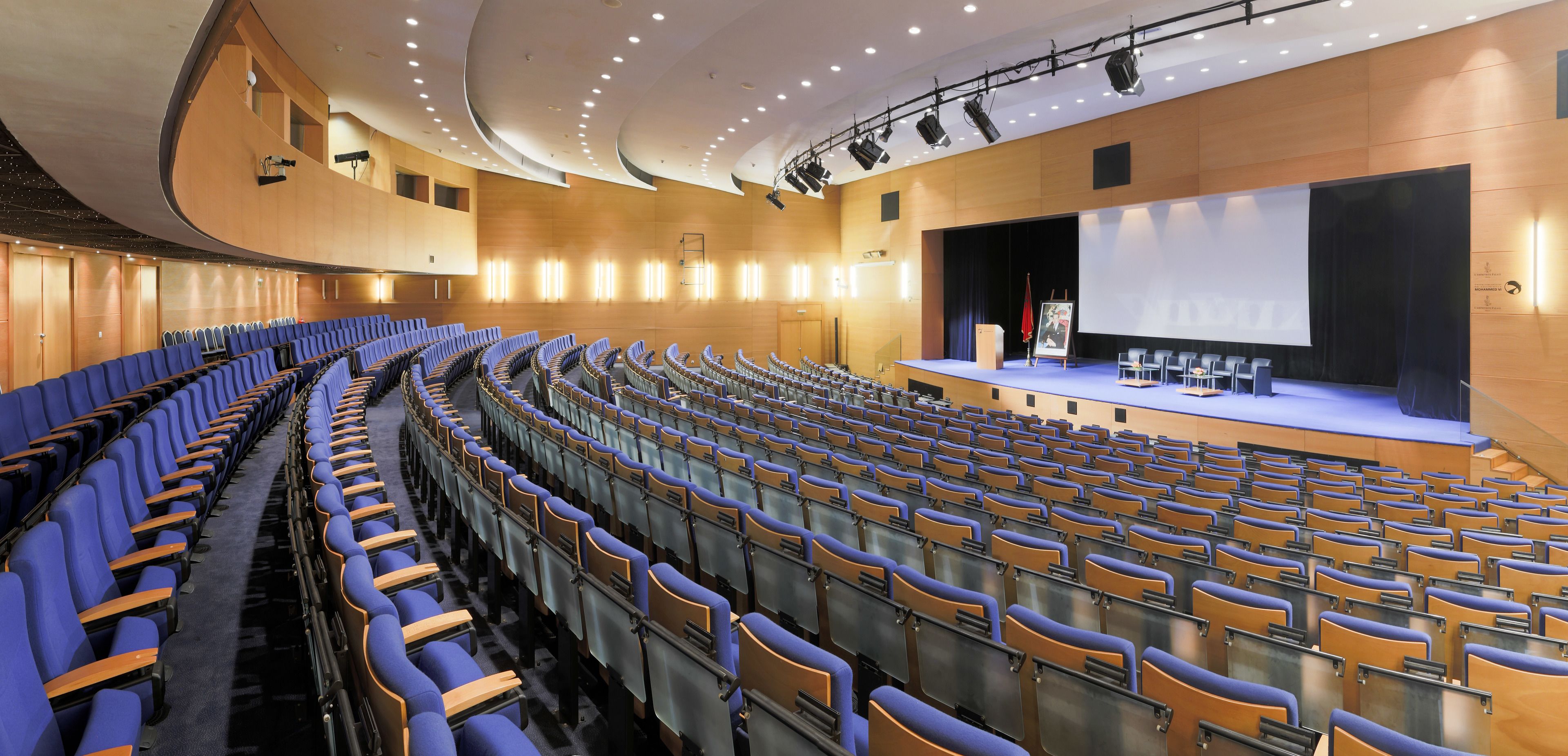 A large auditorium with rows of blue and orange chairs, a stage with a screen, and a curved ceiling with recessed lighting.