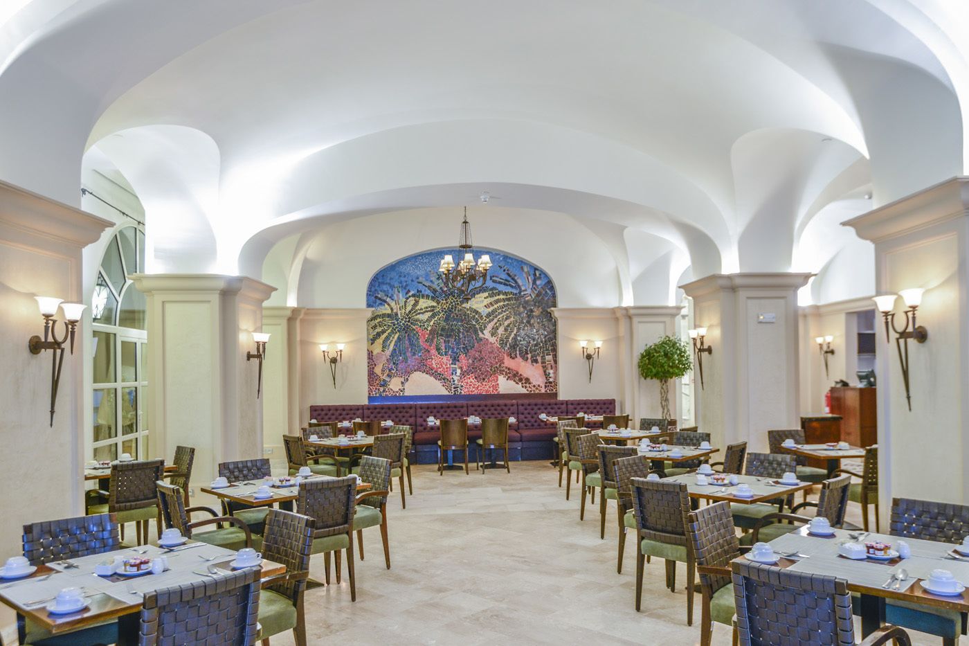 A spacious dining area with high ceilings and a central mural. Tables are set with chairs, and the room has a classic architectural style.