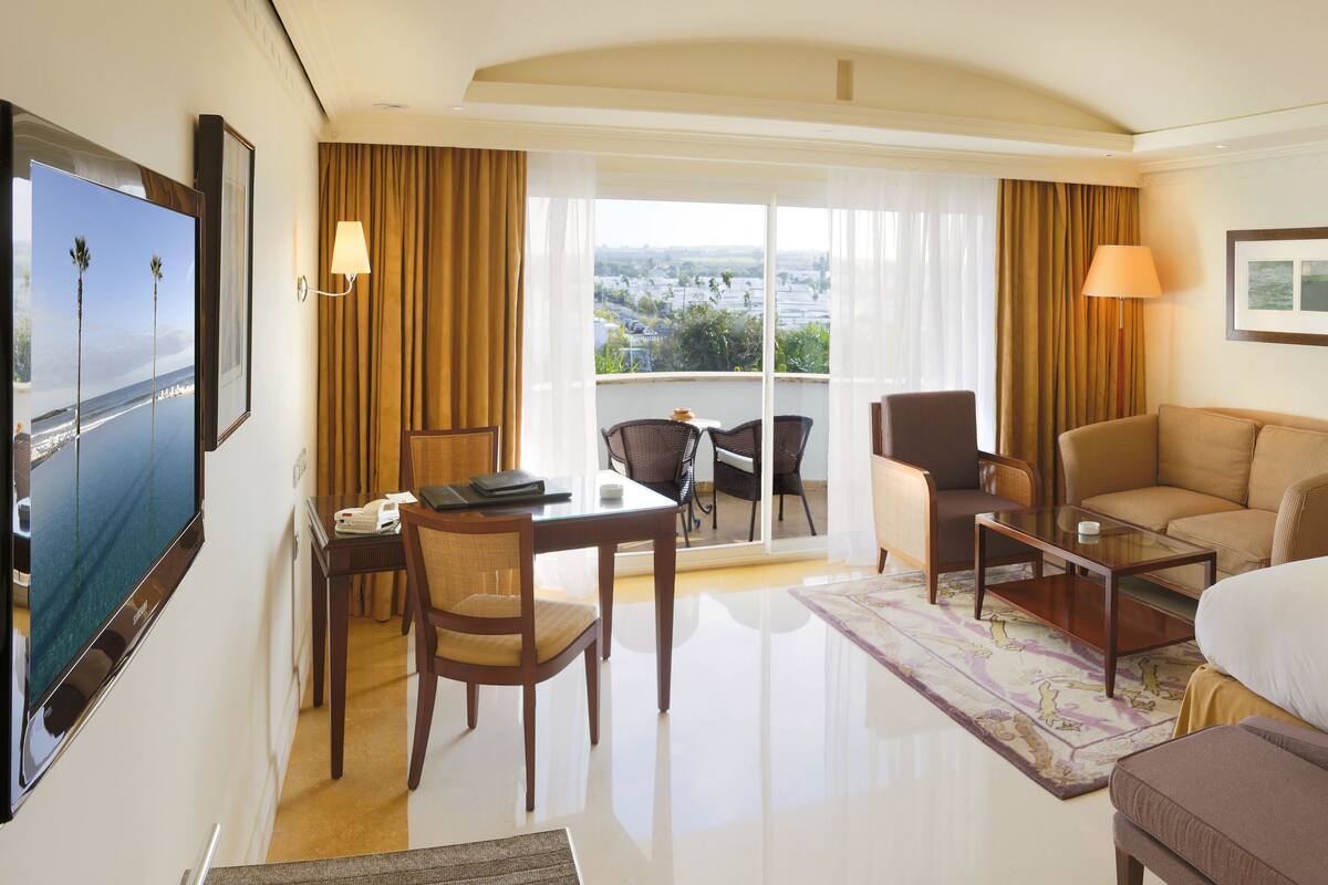 A hotel room with a bed, a desk with a chair, a sofa, a balcony with a view, and a TV on the wall.