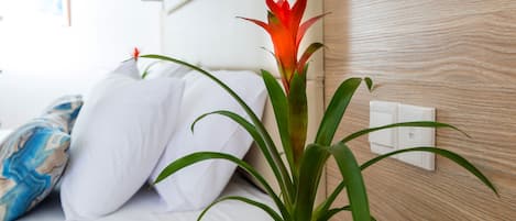 Eine Pflanze in einem Holztopf auf einem Nachttisch neben einem weißen Bett mit blauem Kissen.