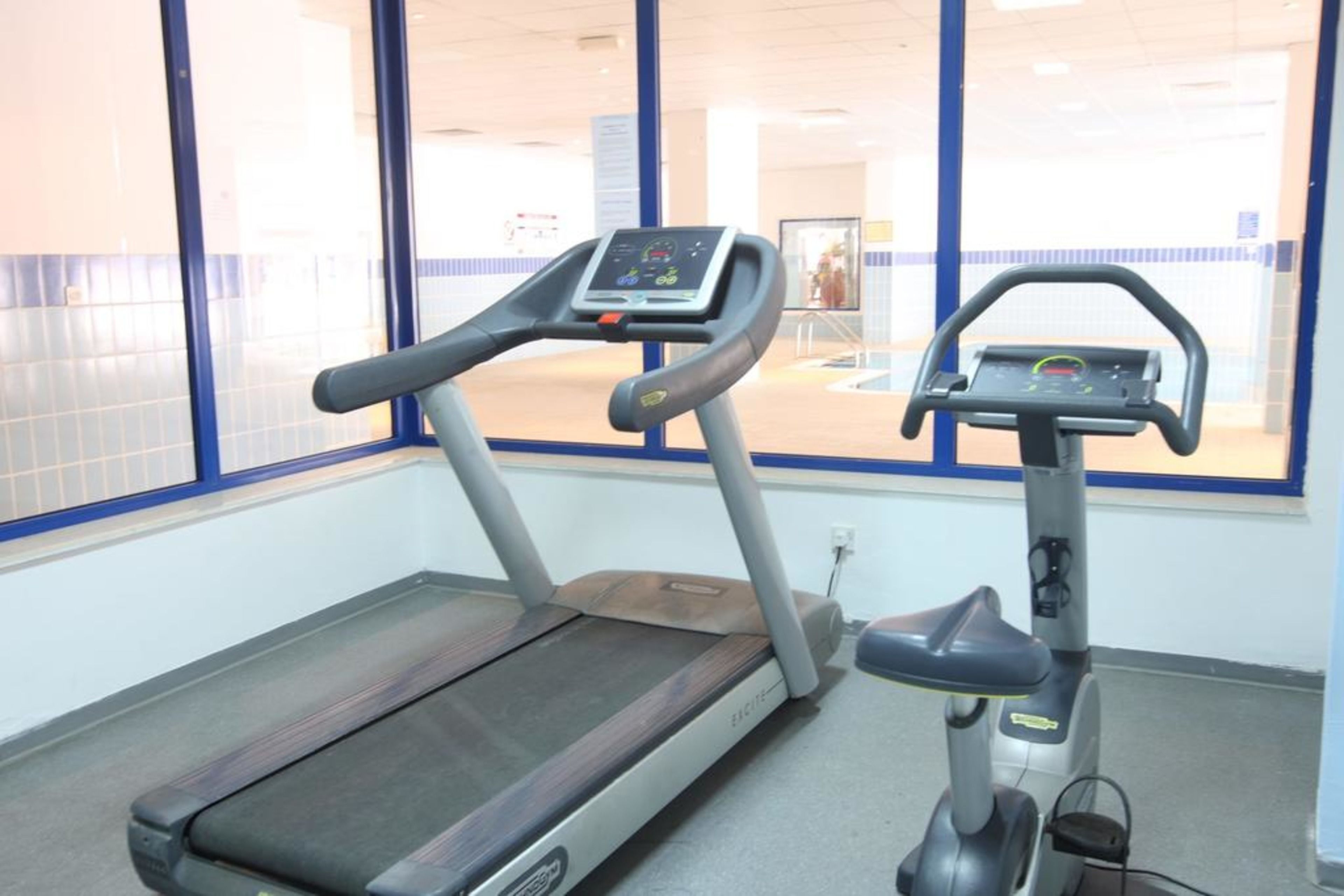 Ein Laufband und ein Crosstrainer in einem Fitnessstudio mit großen Fenstern und Fliesenboden.