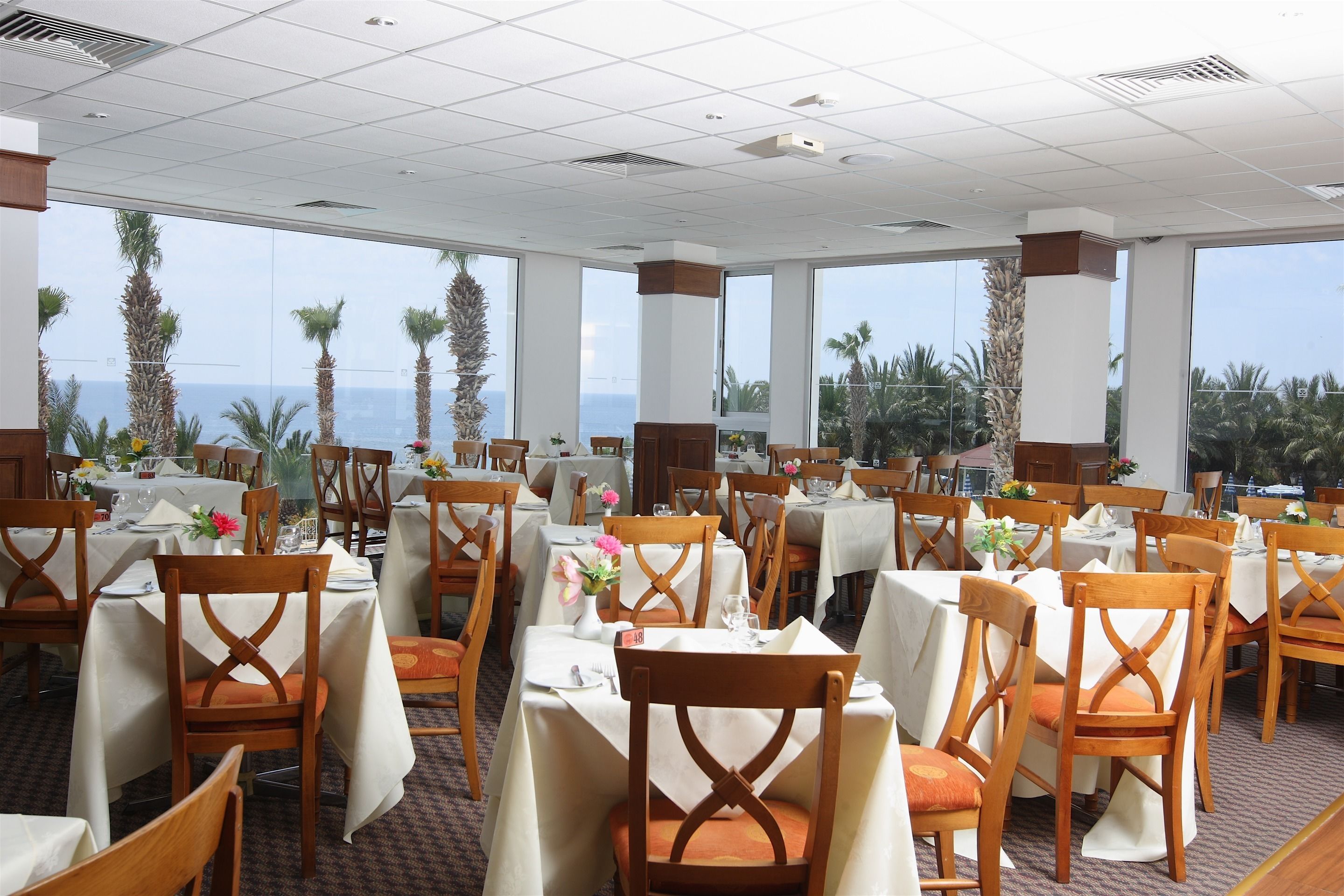 Un ristorante con tavoli apparecchiati, ampie finestre che offrono vista su palme e oceano, e sedie di legno con cuscini arancioni.