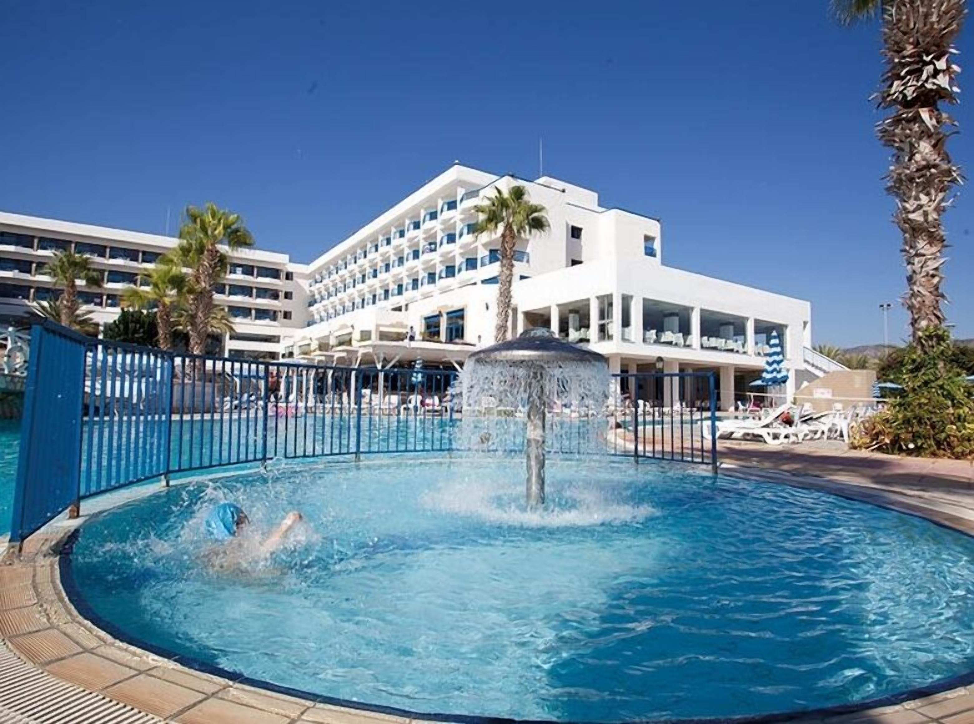 A swimming pool with a water fountain, surrounded by palm trees and a hotel building in the background.