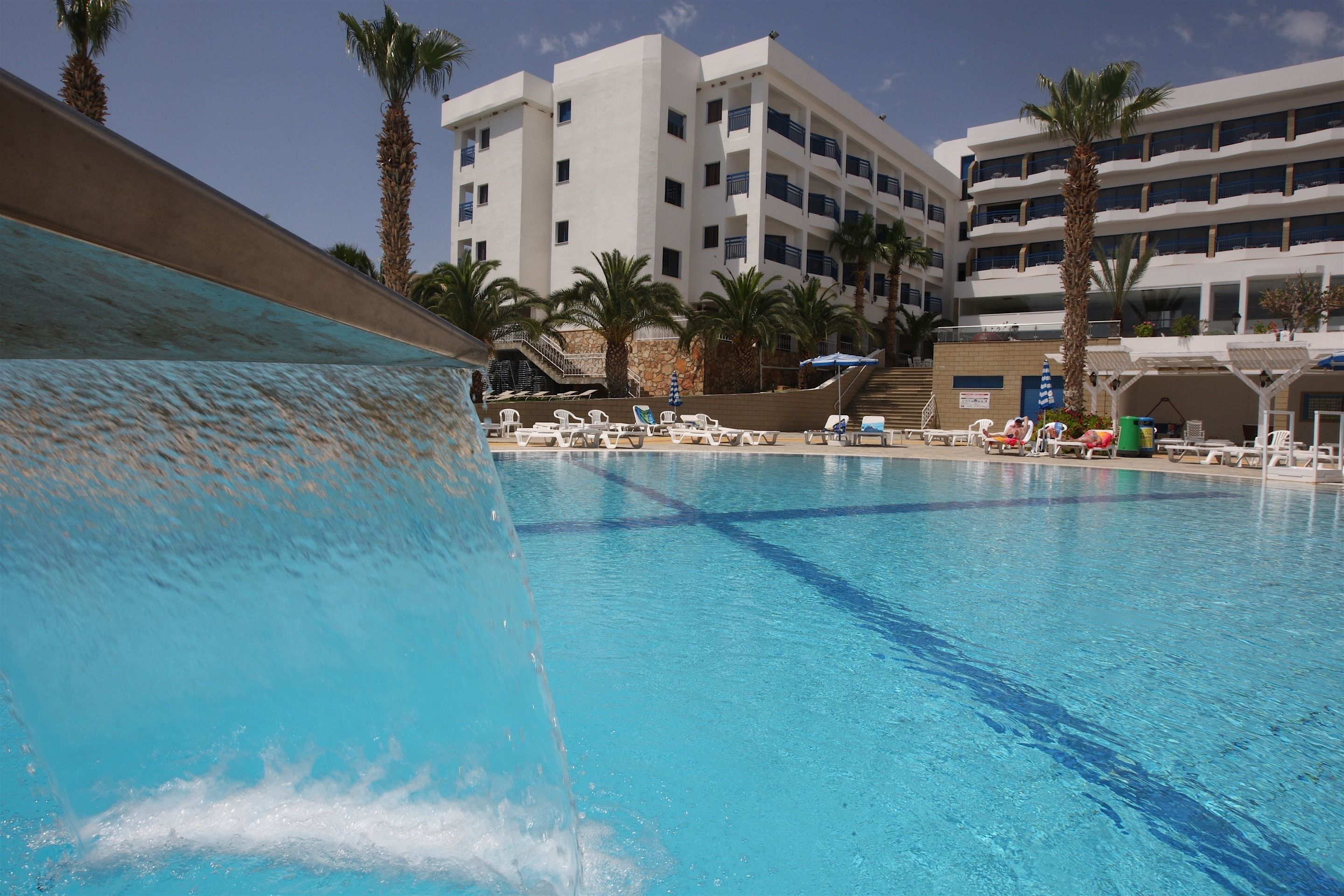 A swimming pool with clear blue water, surrounded by lounge chairs and palm trees, with a multi-story building in the background.