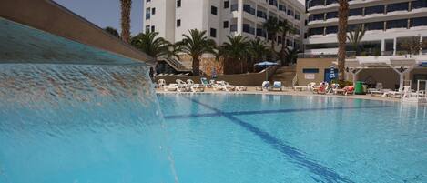 A swimming pool with clear blue water, surrounded by lounge chairs and palm trees, with a multi-story building in the background.