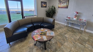 Un área de descanso moderna con un sofá de cuero negro curvo, una mesa de centro redonda con revistas, una planta en maceta y una pequeña mesa con objetos decorativos.