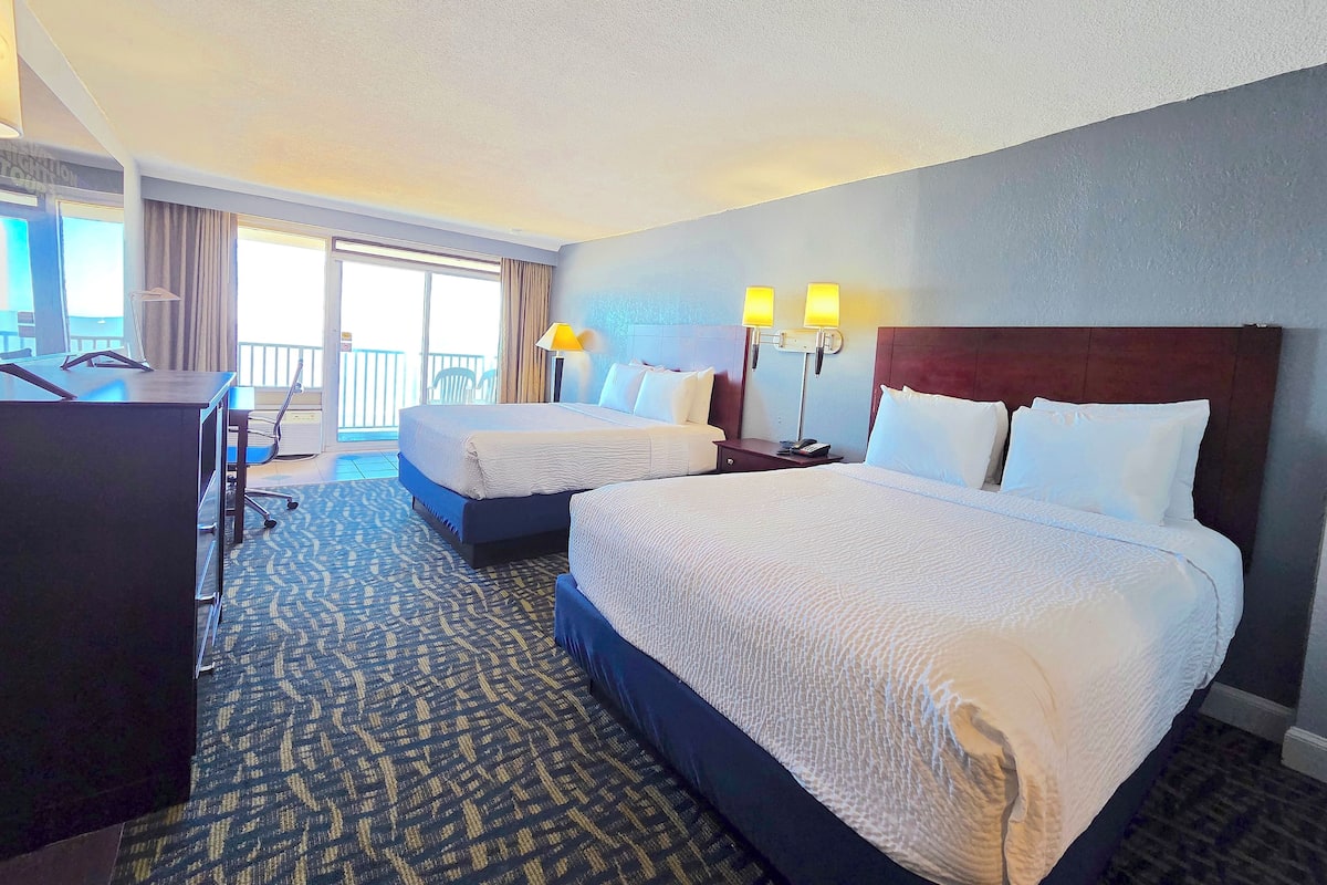A hotel room with two beds, a desk, and a chair. There is a balcony with a view.