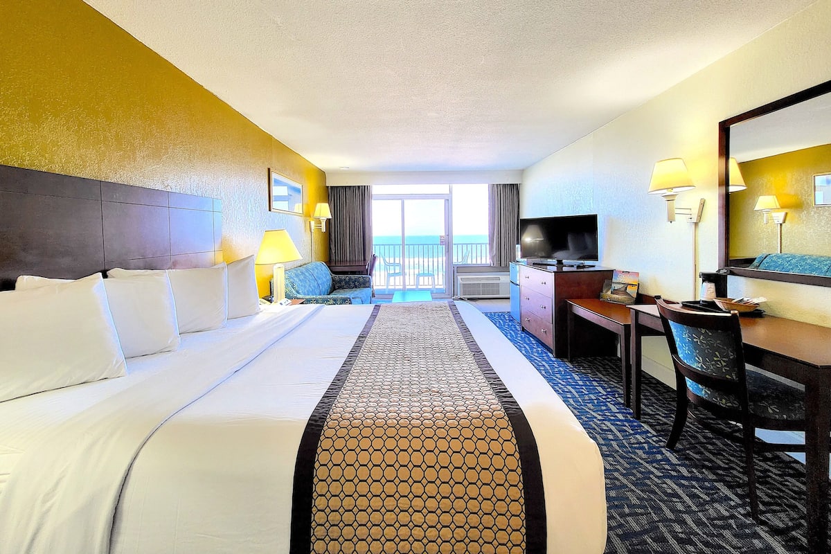 A hotel room with a large bed, a desk with a computer, a TV, and a view of the ocean through the balcony doors.