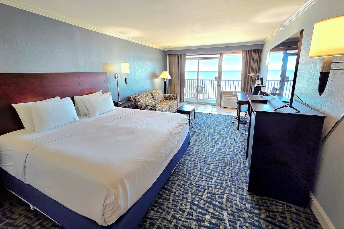 A hotel room with a large bed, a desk, a chair, and a view of the sea through a sliding door.