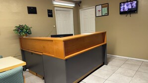 A reception desk with a wooden front and grey paneling, a potted plant, a closed door, and a security camera on the wall. - Americas Best Value Inn Wendover (Wendover)