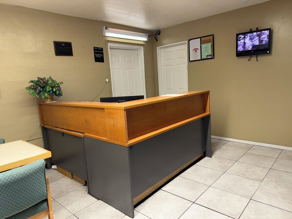 A reception desk with a wooden front and grey paneling, a potted plant, a closed door, and a security camera on the wall.