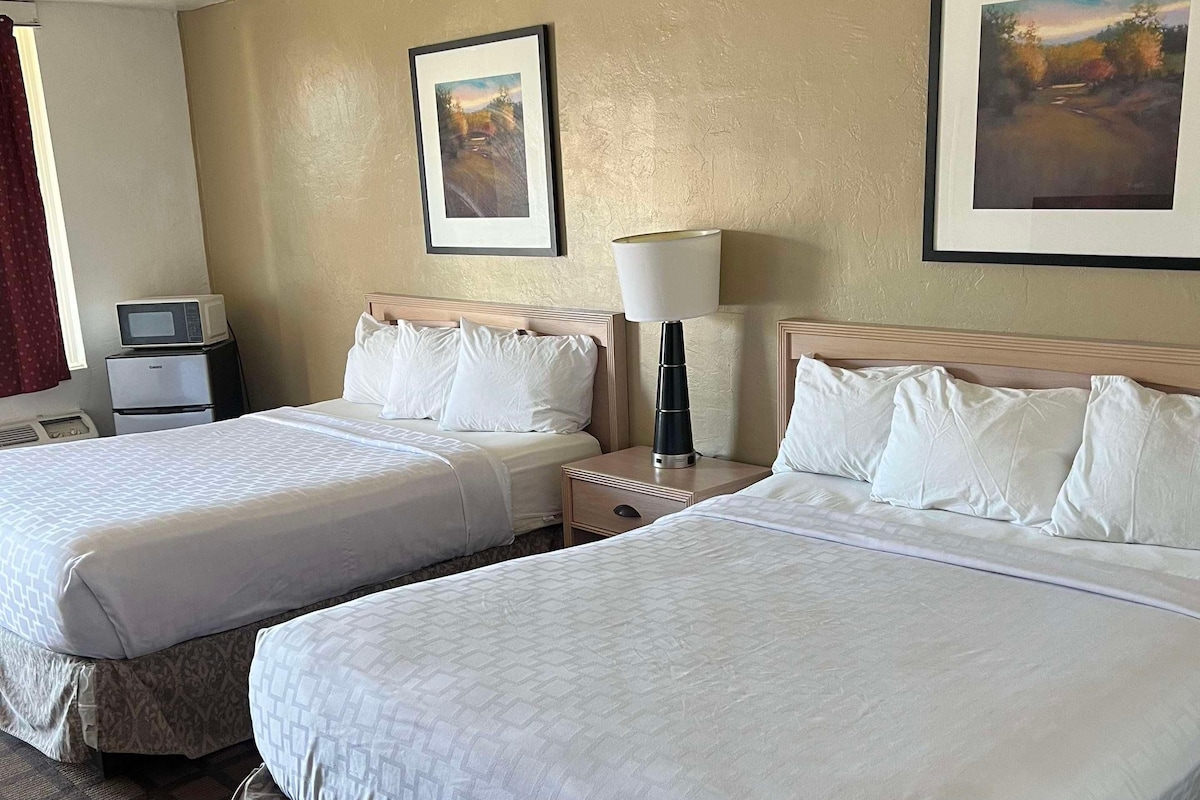 A hotel room with two beds, a bedside table, a lamp, a microwave, and two framed pictures on the wall.