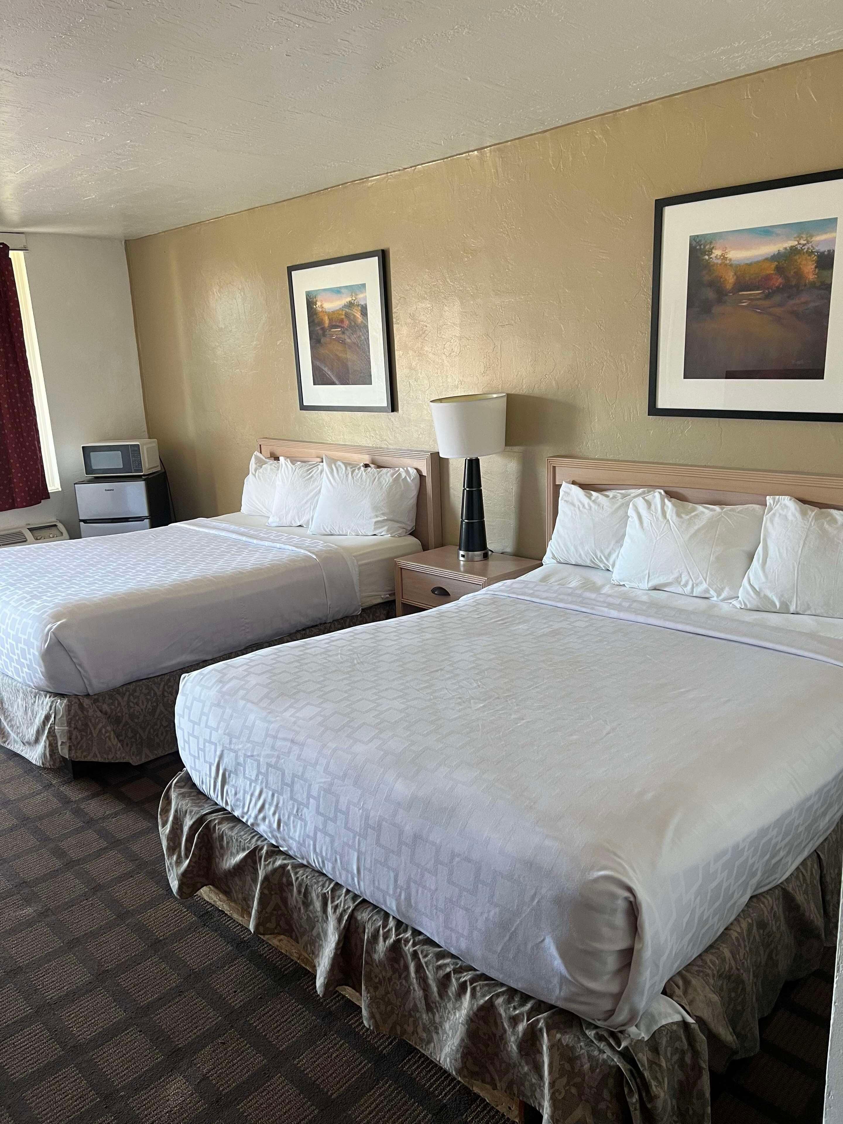 Una habitación de hotel con dos camas, una mesita de noche, una lámpara, un microondas y dos cuadros enmarcados en la pared.