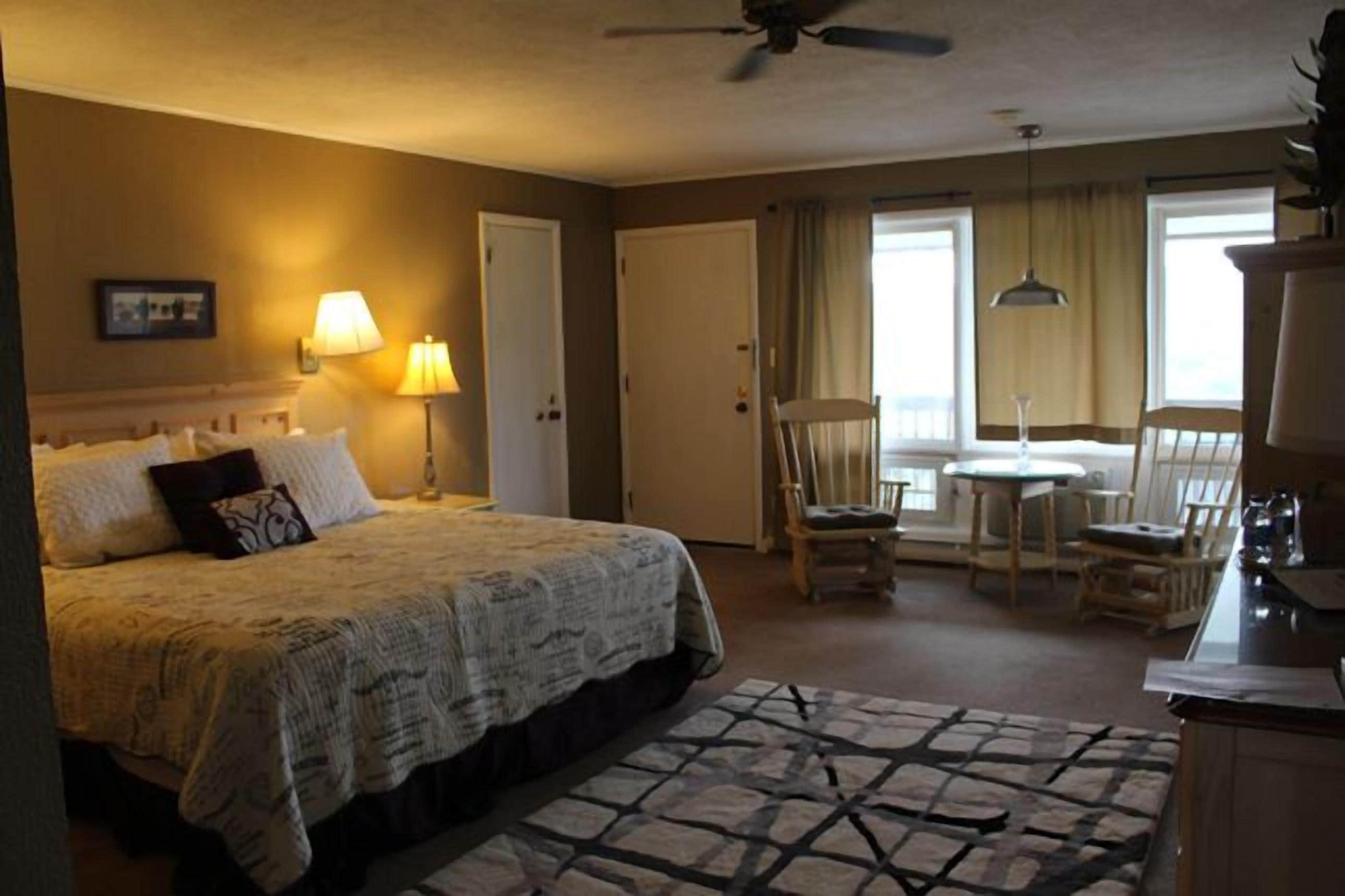A hotel room with a bed, a desk, a chair, a lamp, a ceiling fan, and a view of the outdoors.