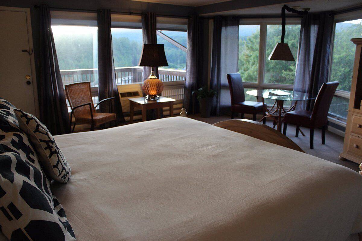 A hotel room with a large bed, a desk with a lamp, two chairs, a small table, and a view of the outdoors through large windows.