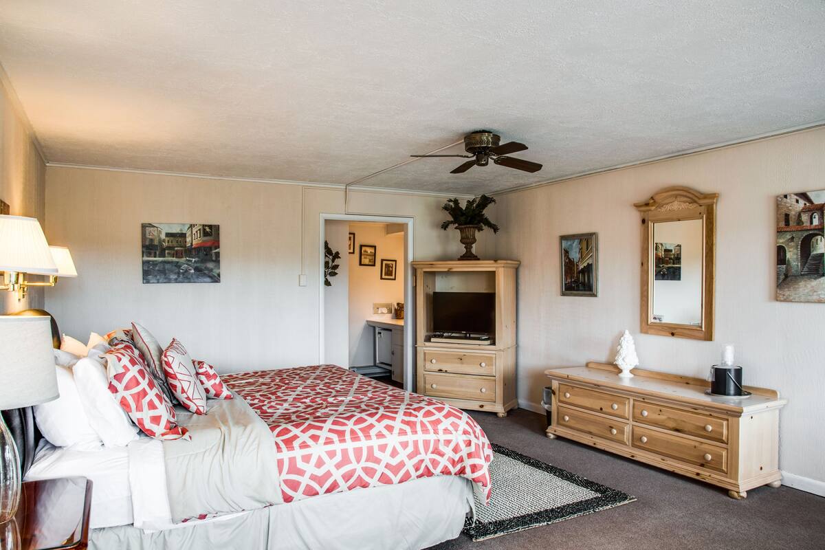A bedroom with a bed, dresser, vanity, ceiling fan, and artwork on the walls.