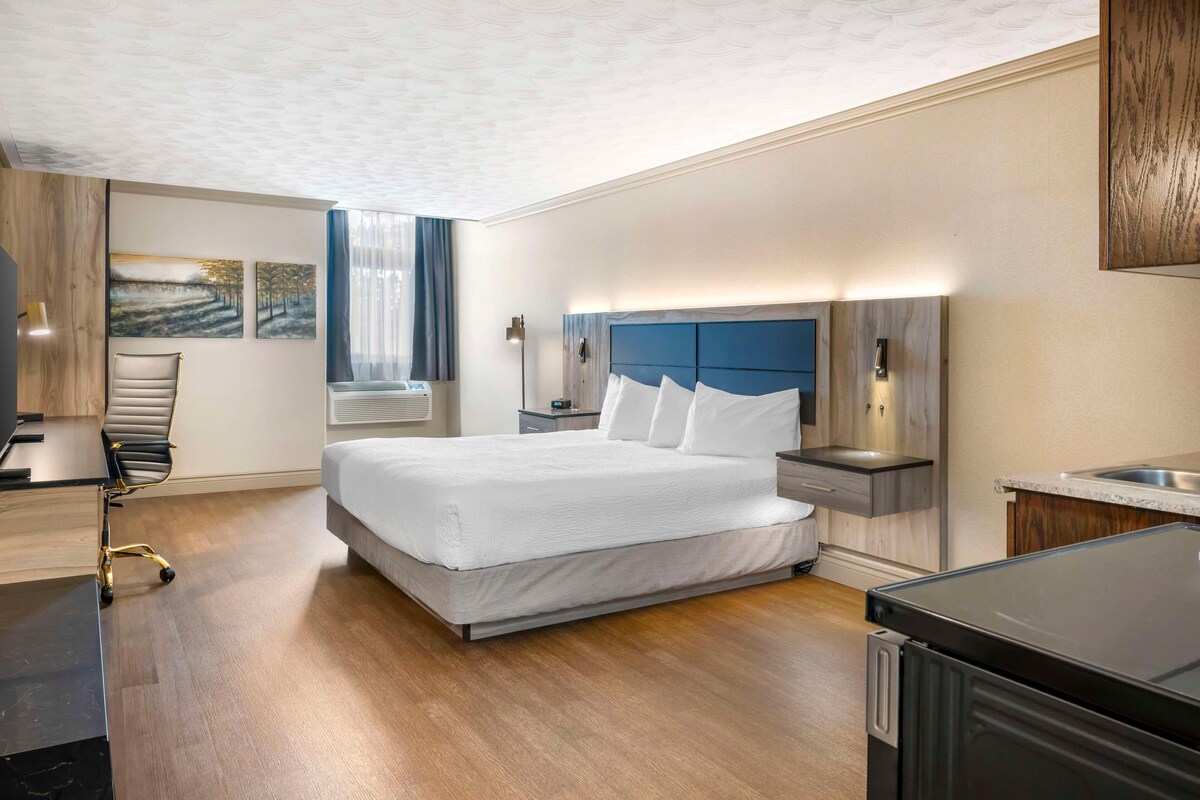 A hotel room with a large bed, bedside tables, a desk with a chair, a wardrobe, and a view of the outdoors.