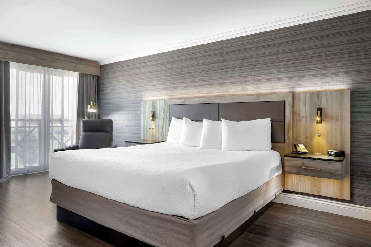 Une chambre d’hôtel moderne avec un grand lit, une tête de lit en bois, des tables de chevet avec des lampes et une vue sur l’extérieur grâce à de grandes fenêtres.