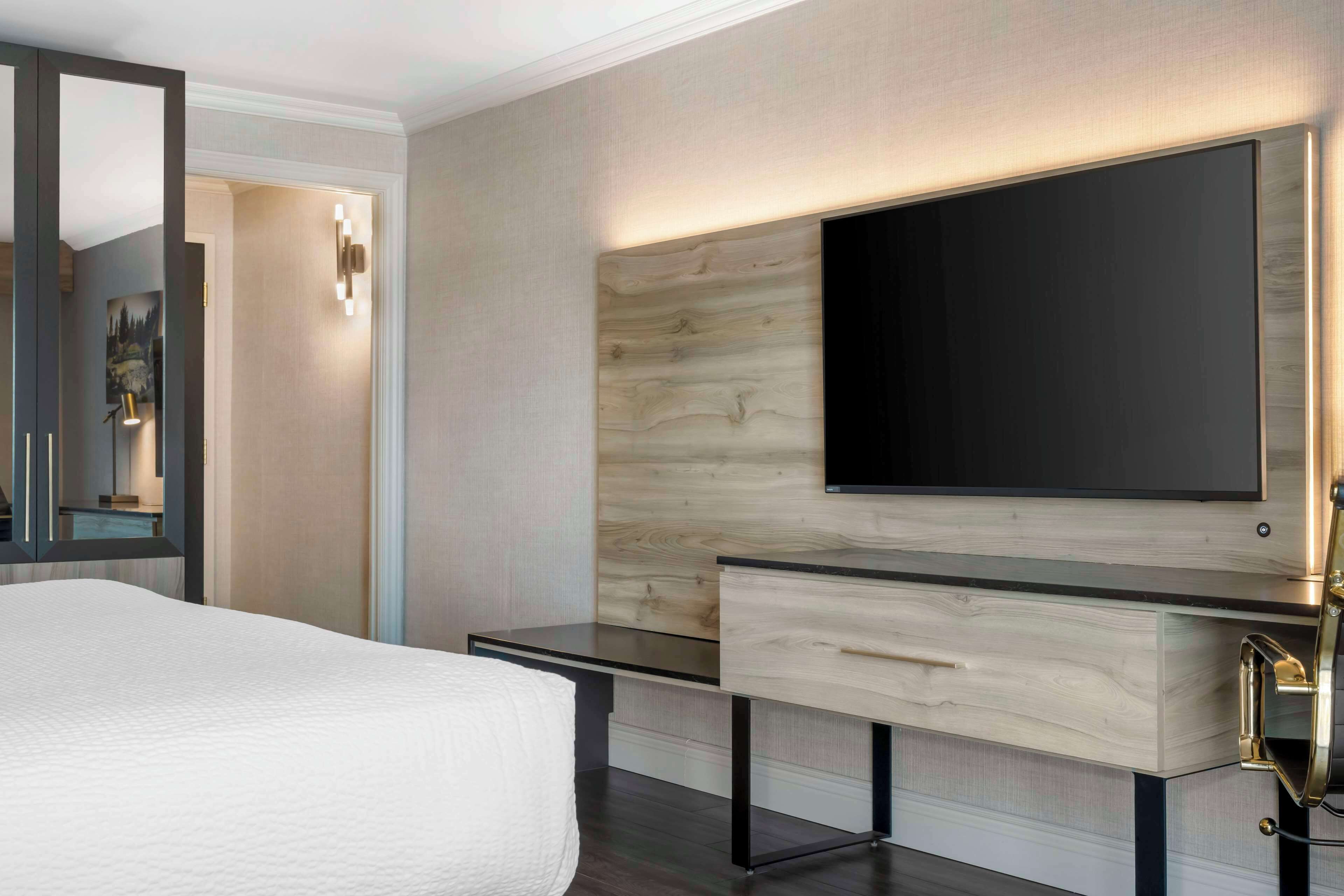 A modern hotel room with a large bed, a wooden accent wall, a flat-screen TV, and a desk with drawers.