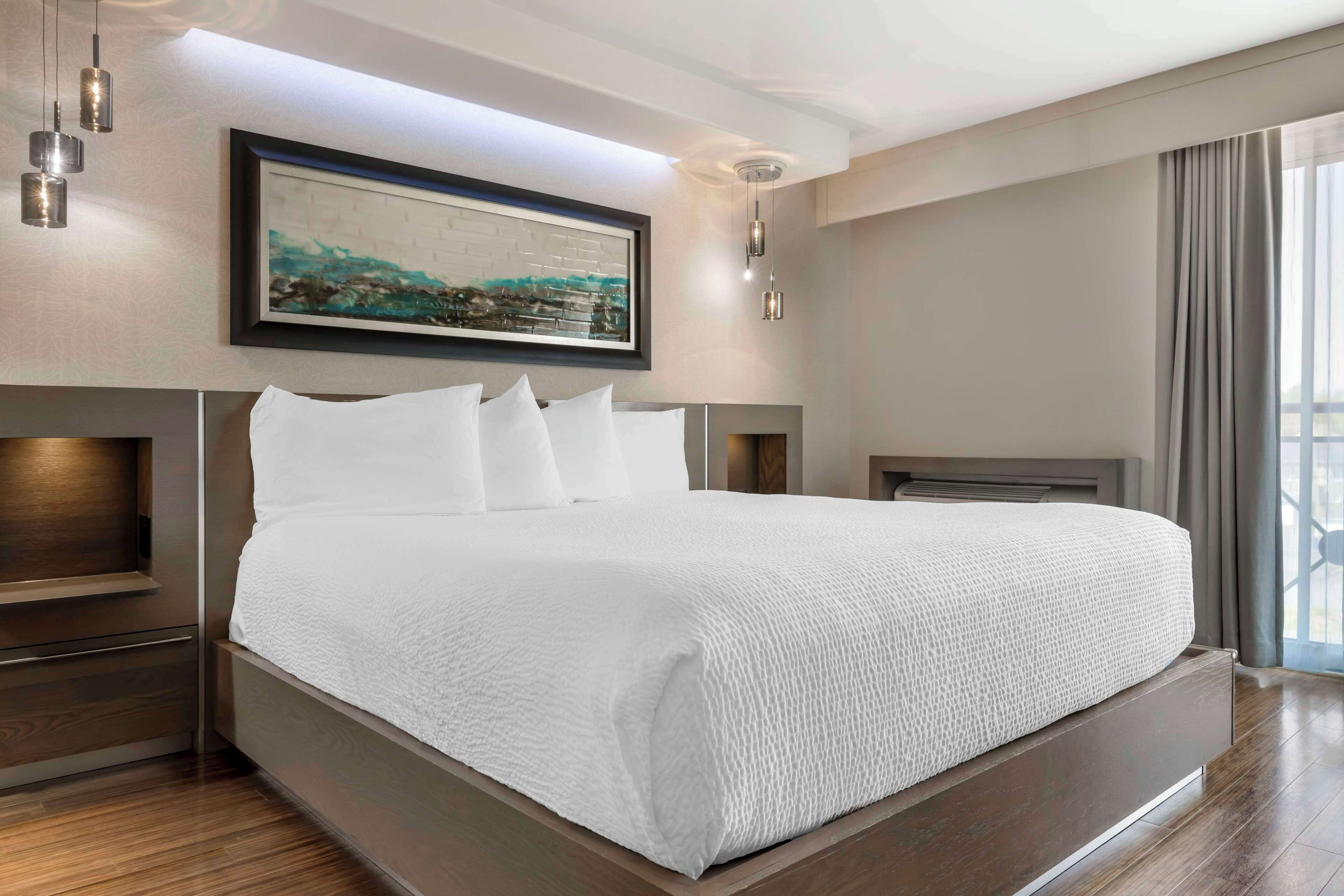 Una habitación de hotel moderna con una cama grande, una obra de arte enmarcada sobre la cama y vistas al exterior a través de la ventana.