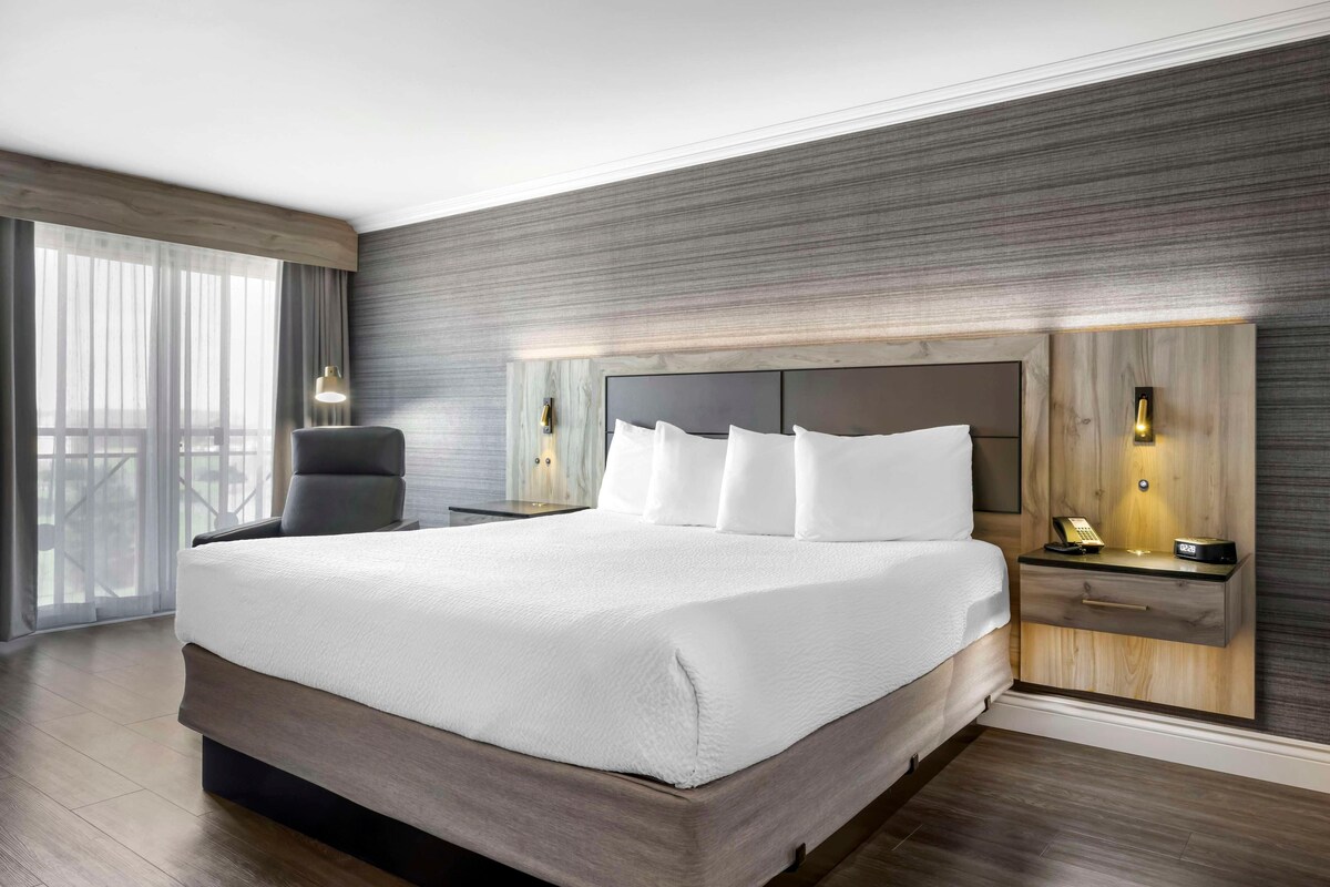 A modern hotel room with a large bed, a wooden headboard, a nightstand with a lamp, and a chair.