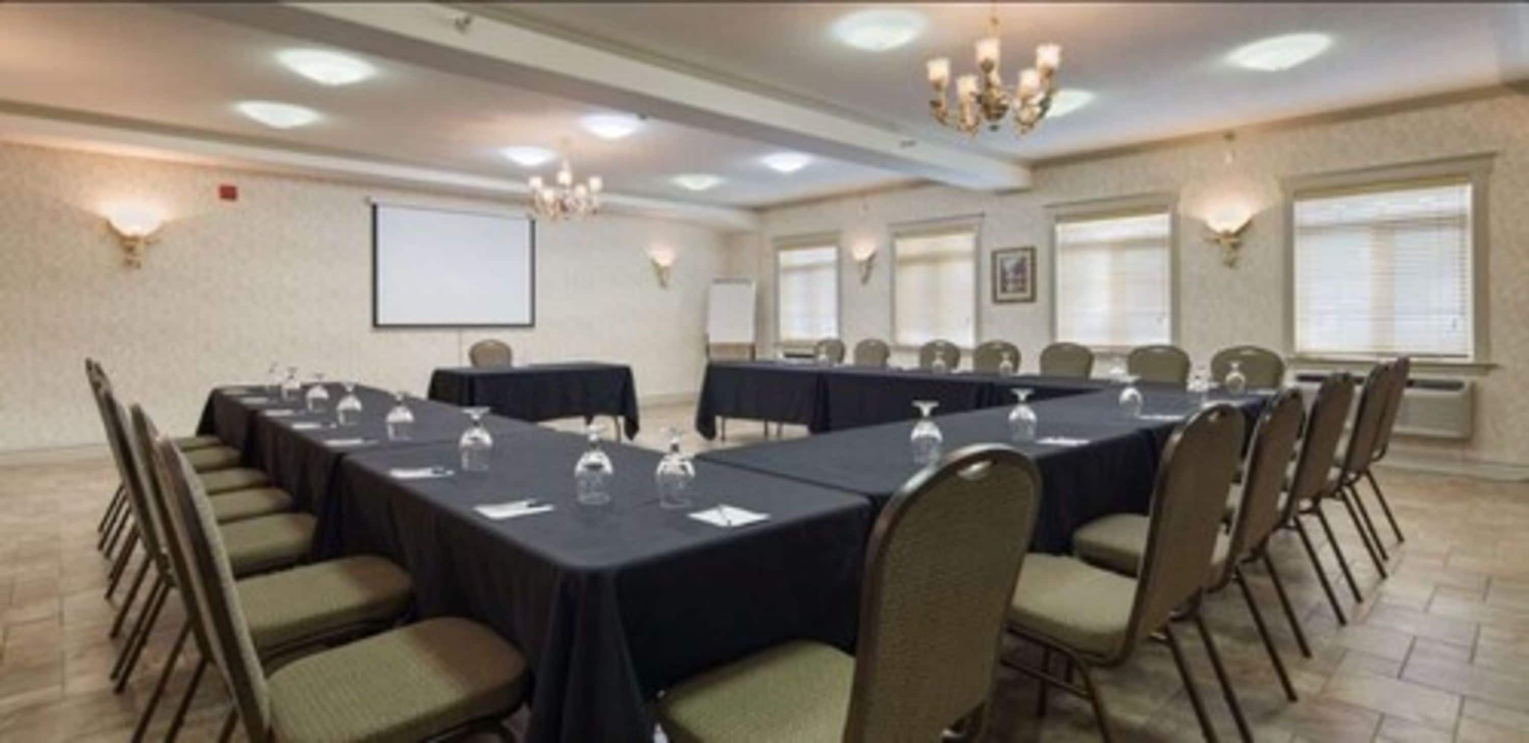 Un salón de conferencias con mesas largas, sillas, una pantalla de proyector y luces empotradas en la pared.