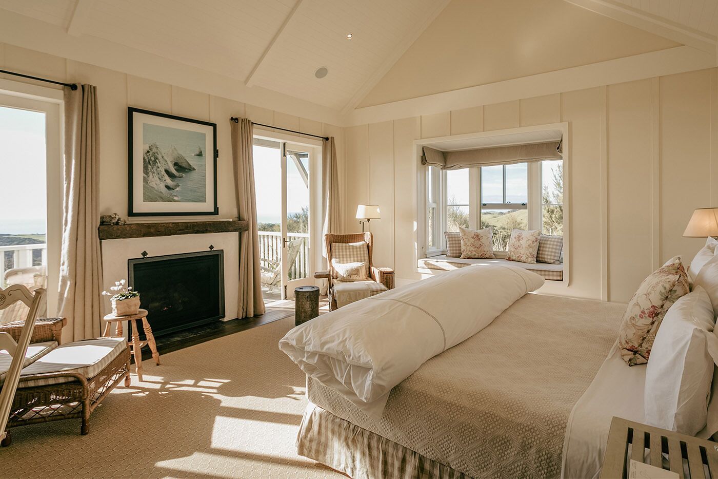 A spacious bedroom with a large bed, a fireplace, a view of the outdoors, and a balcony with seating.