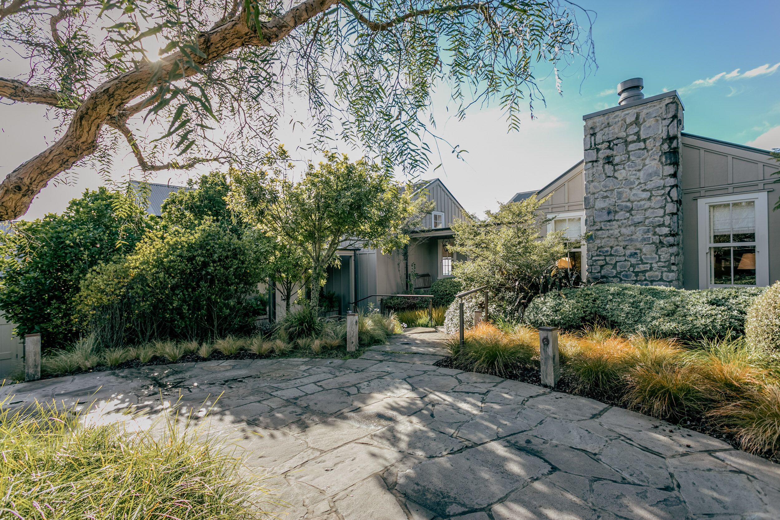A stone pathway leading to a modern house with a stone chimney, surrounded by lush greenery and a large tree.