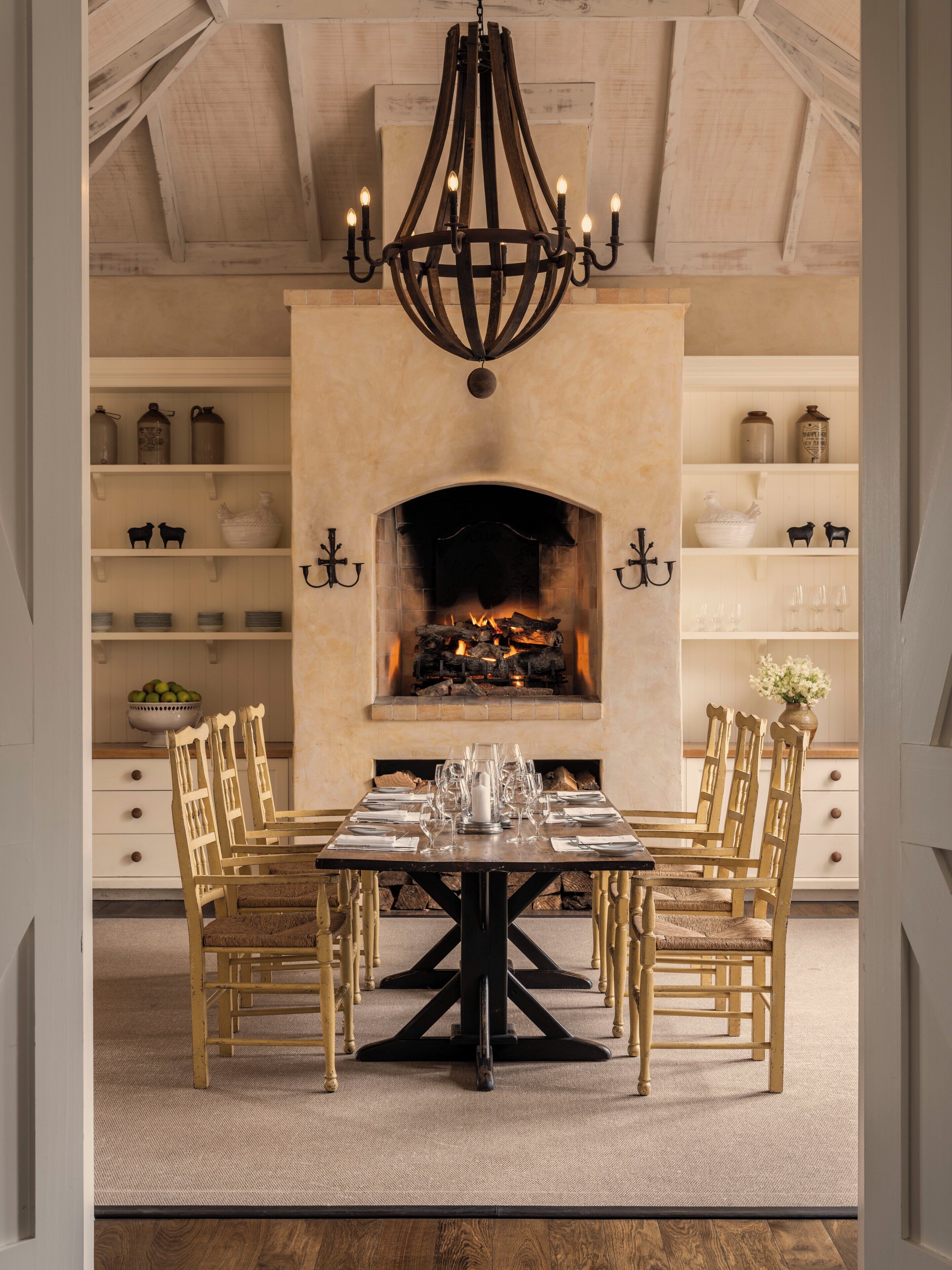 A dining room with a fireplace, wooden chairs, a table set for six, and shelves with kitchenware.