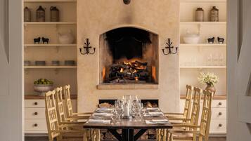 A dining room with a fireplace, wooden chairs, a table set for six, and shelves with kitchenware.
