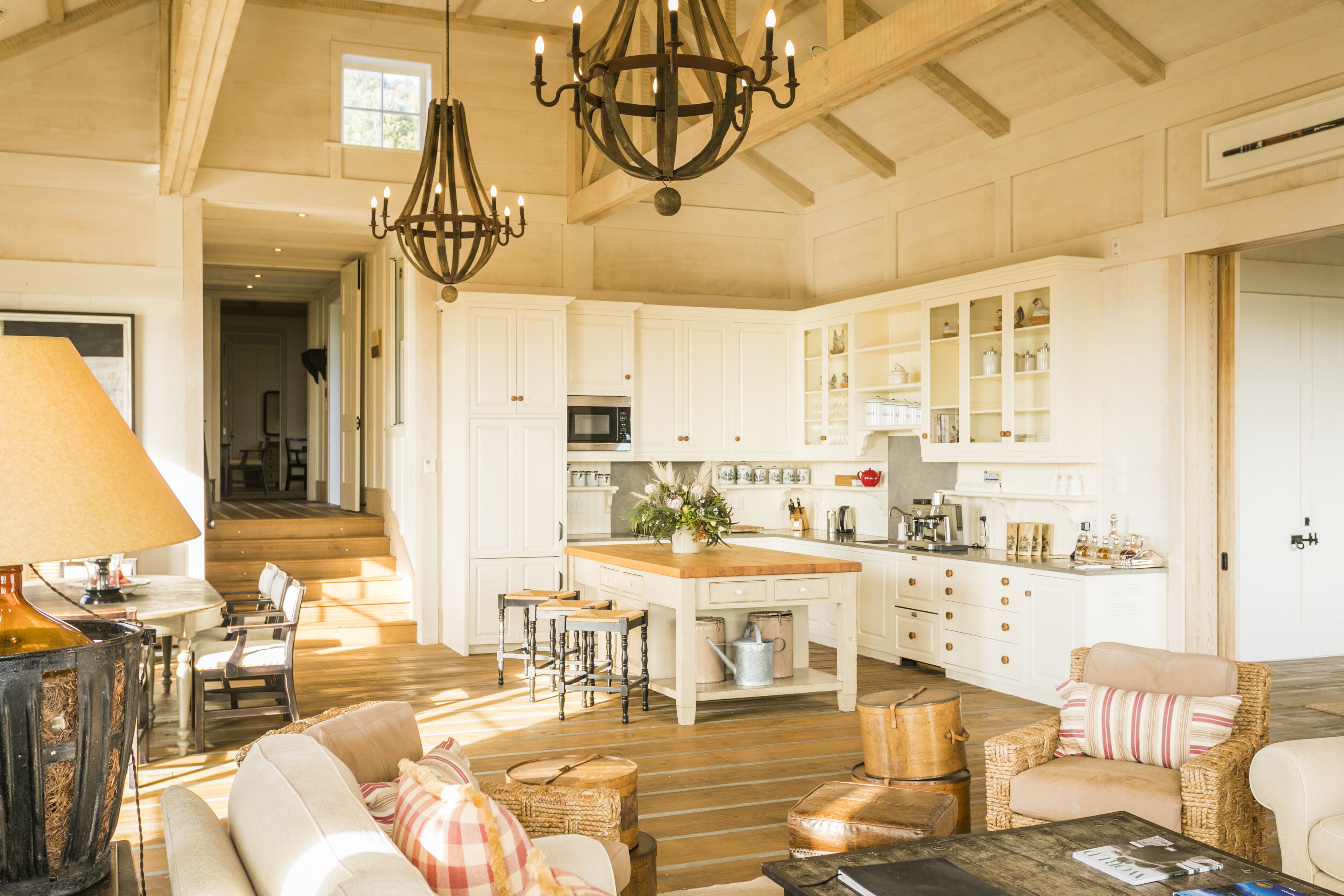 A spacious kitchen with white cabinetry, a central island, and a chandelier. There are wooden floors, a dining area with chairs, and a living room with sofas and a coffee table.