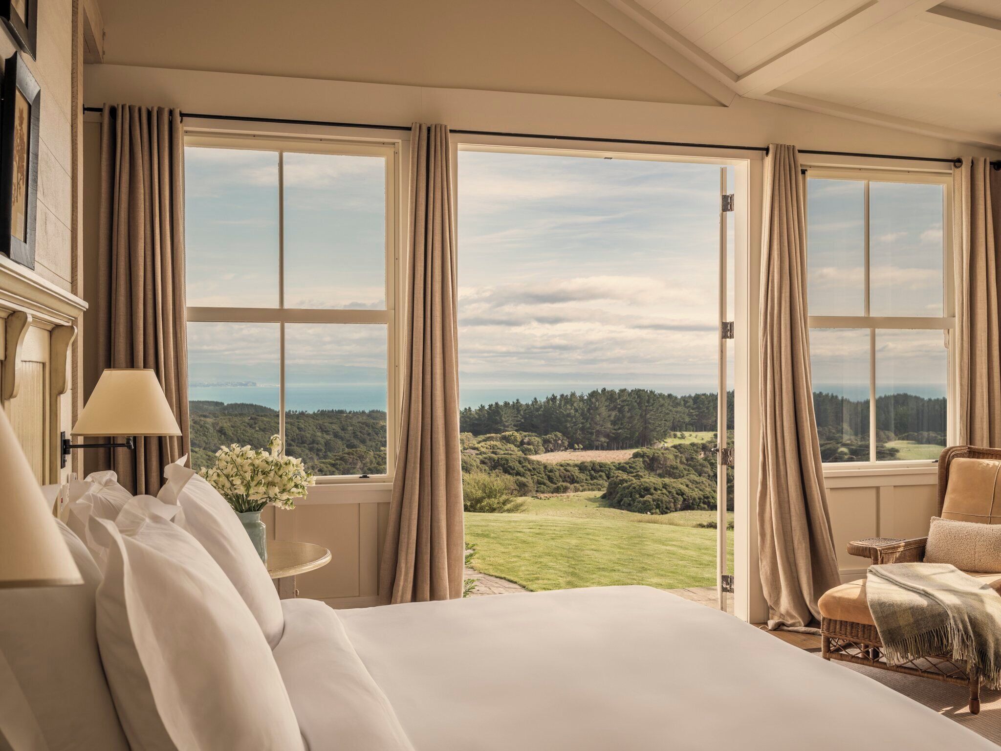 Une chambre d’hôtel avec un grand lit, une chaise, une table et une vue sur un paysage verdoyant, visible à travers de grandes fenêtres.