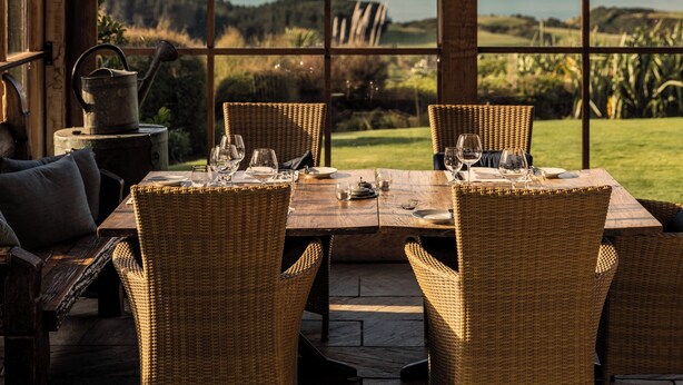 Una mesa de madera para comer con sillas de mimbre, dispuesta para cuatro personas, con vistas a un paisaje pintoresco que incluye el mar y colinas.