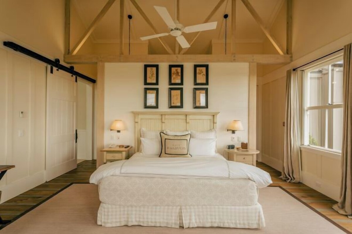 A bedroom with a large bed, wooden ceiling beams, a ceiling fan, framed artwork on the wall, and bedside lamps.